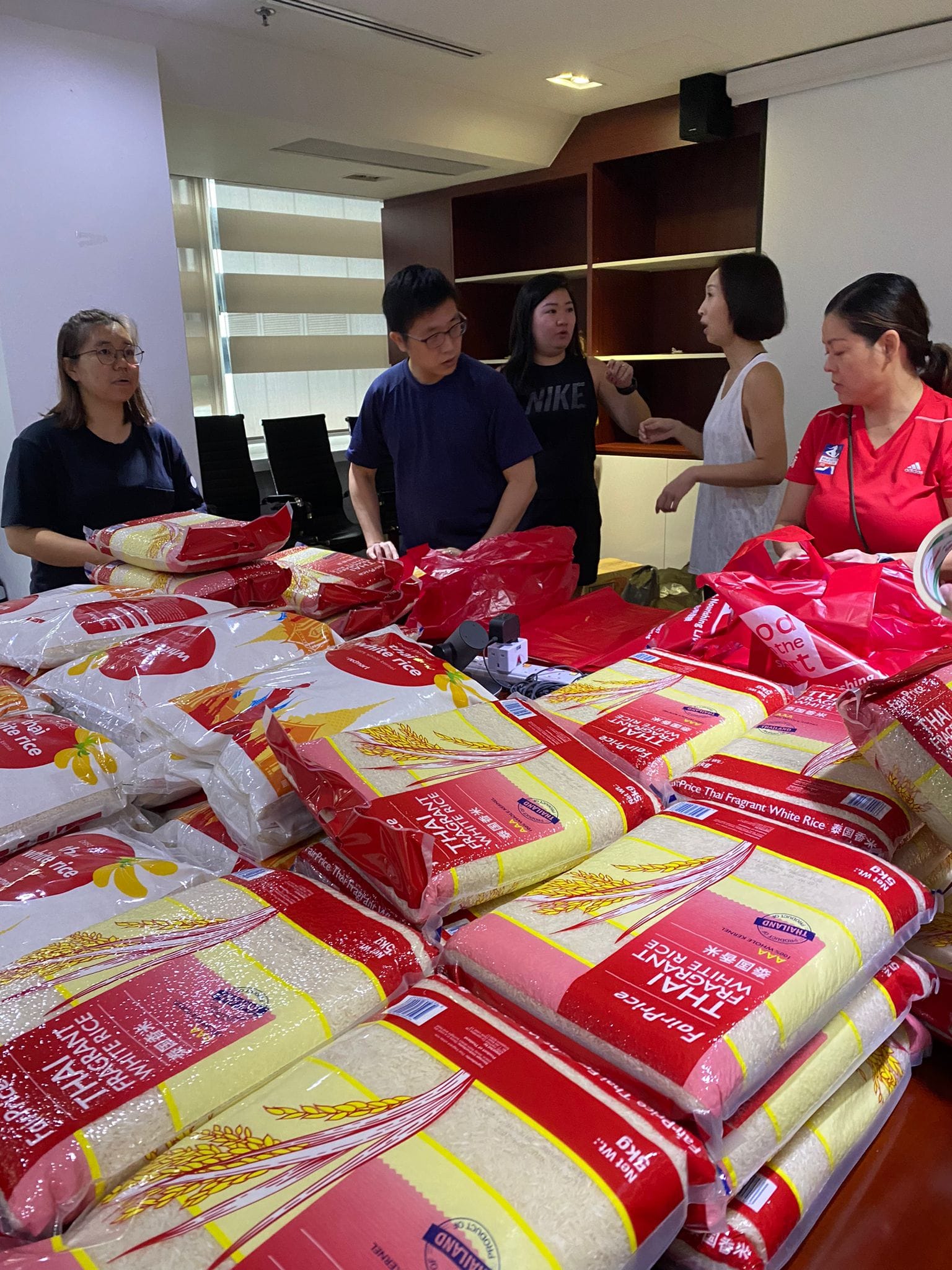 Five people stand around a table stacked with many large bags of Thai Fragrant White Rice in an indoor setting.